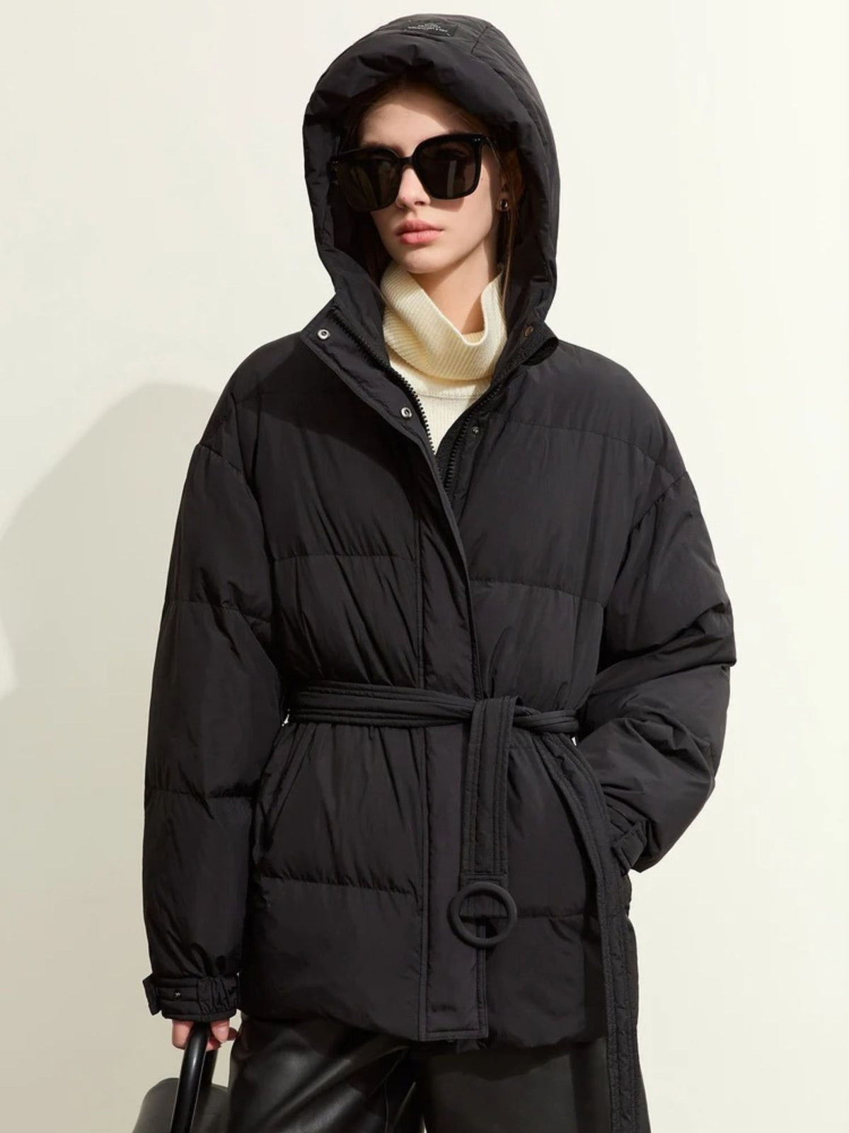 Model wearing black belted puffer jacket with hood up and sunglasses